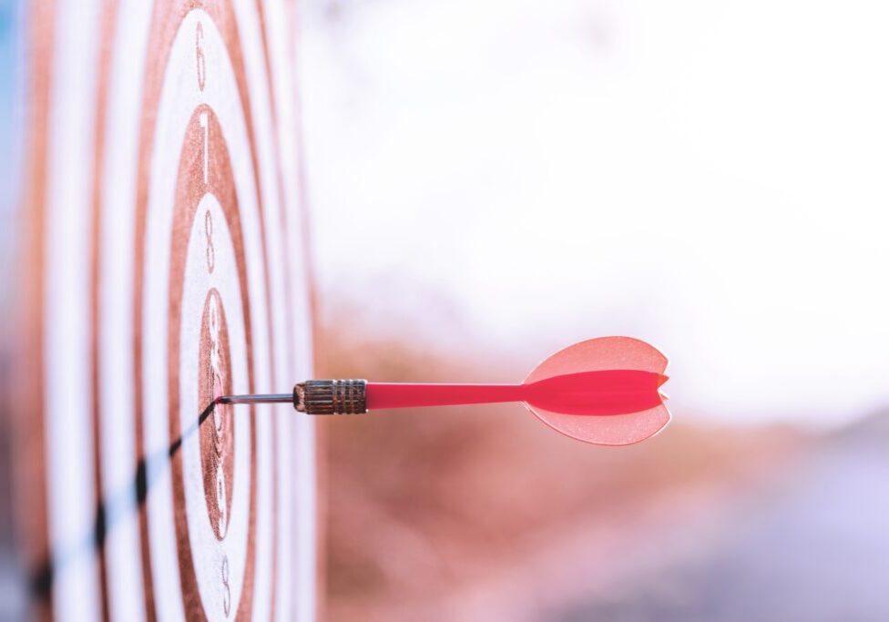 Close up red dart arrow hitting target center dartboard on sunset background. Business targeting and focus concept.