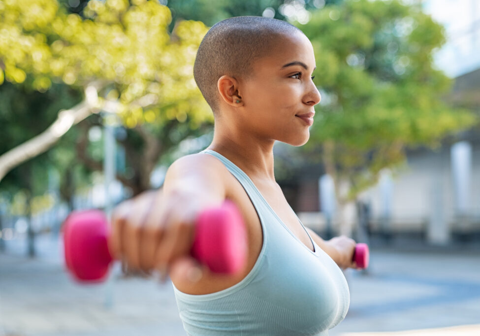 Active curvy woman using dumbbells in the park. Side view of young woman doing stretching exercise using dumbbells. Active athlete working out on fitness routine to lose weight.