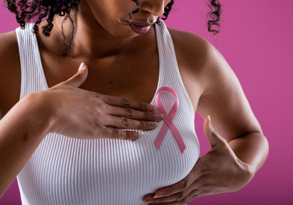 Midsection of african american mid adult woman with pink breast cancer awareness ribbon on breast. pink background, breast, cancer, medical, ribbon, awareness, support, healthcare and alertness.